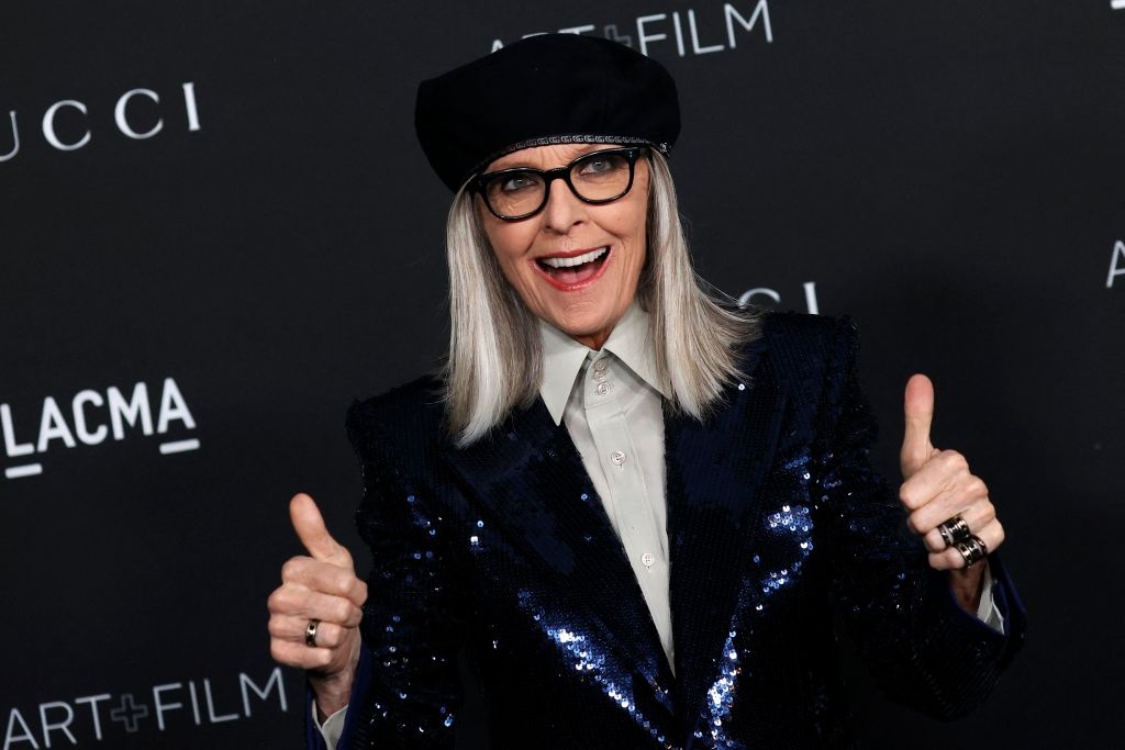 Person on red carpet in a sequin jacket, beret, and glasses, smiling and giving two thumbs up in front of a backdrop with brand logos