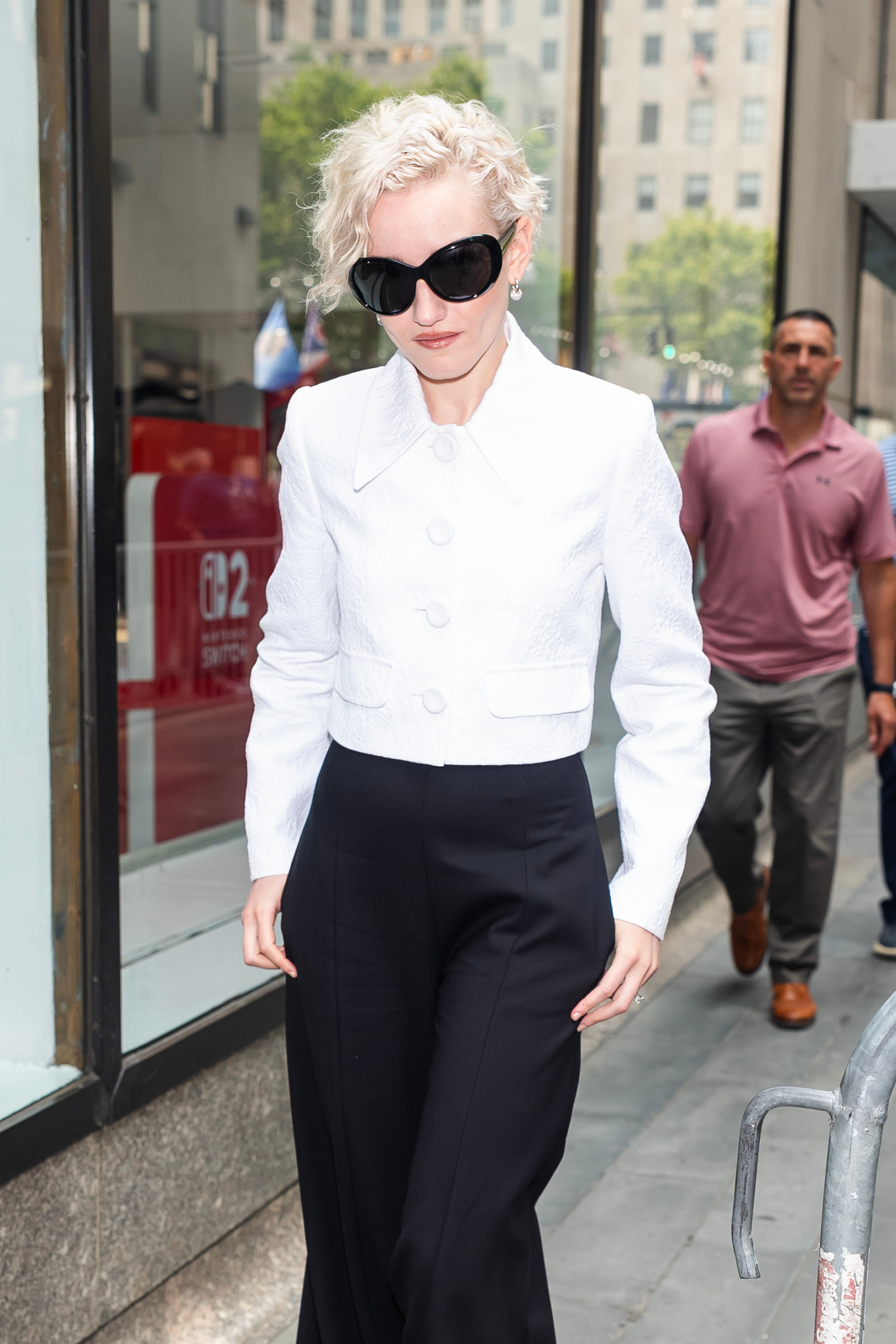 Julia Garner wearing a tailored jacket and pants walks outside, accessorized with large sunglasses