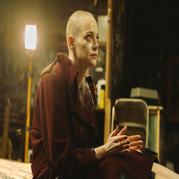 A person with a shaved head and dramatic makeup sits on a bed in a dimly lit room, wearing a long coat, hands clasped together