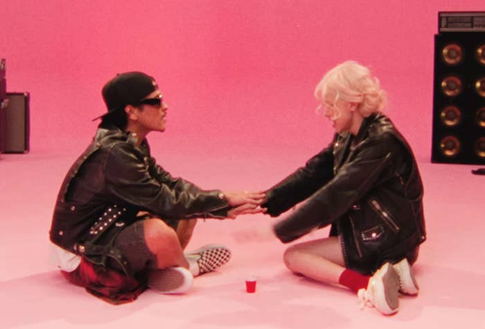 Bruno and Rosé sitting and facing each other with their hands stacked, playing a drinking game