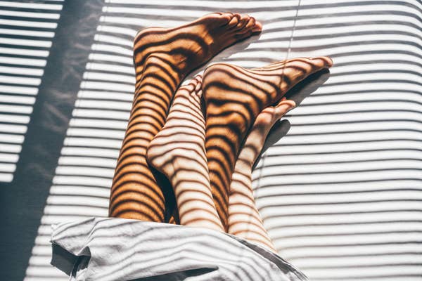 Legs intertwined under sunlight filtered through blinds, creating striped shadows on skin. Sheets partially cover the feet, enhancing the intimate scene