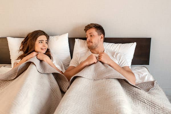 A couple lies in bed, looking at each other with soft expressions, under a shared quilt