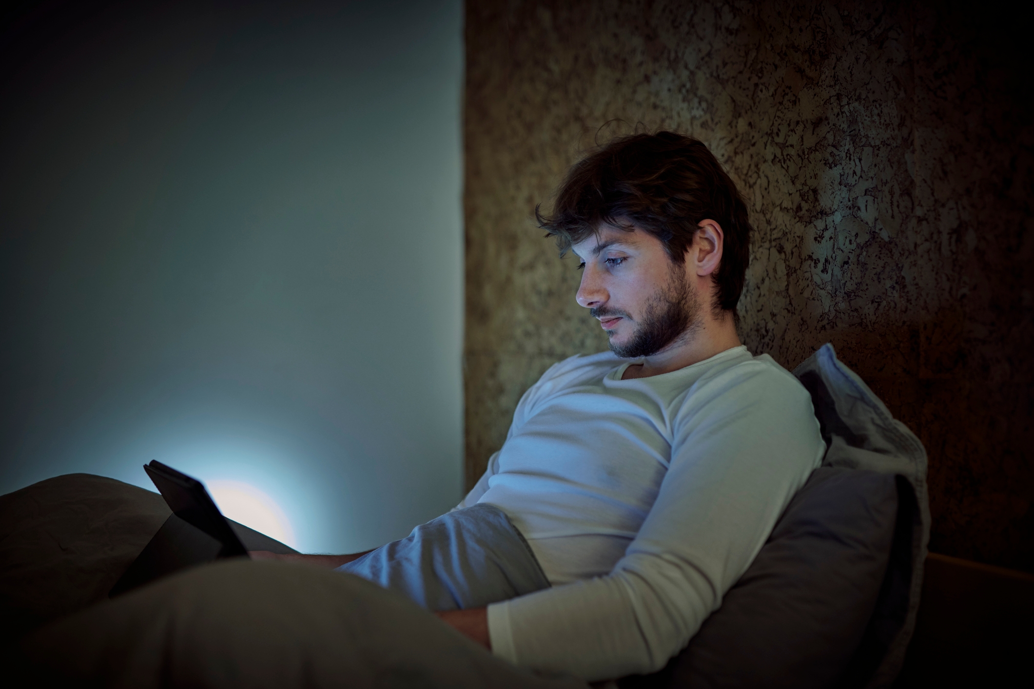 A person in bed, propped up with pillows, attentively reads a tablet screen in a dimly lit room, suggesting a relaxed or intimate setting