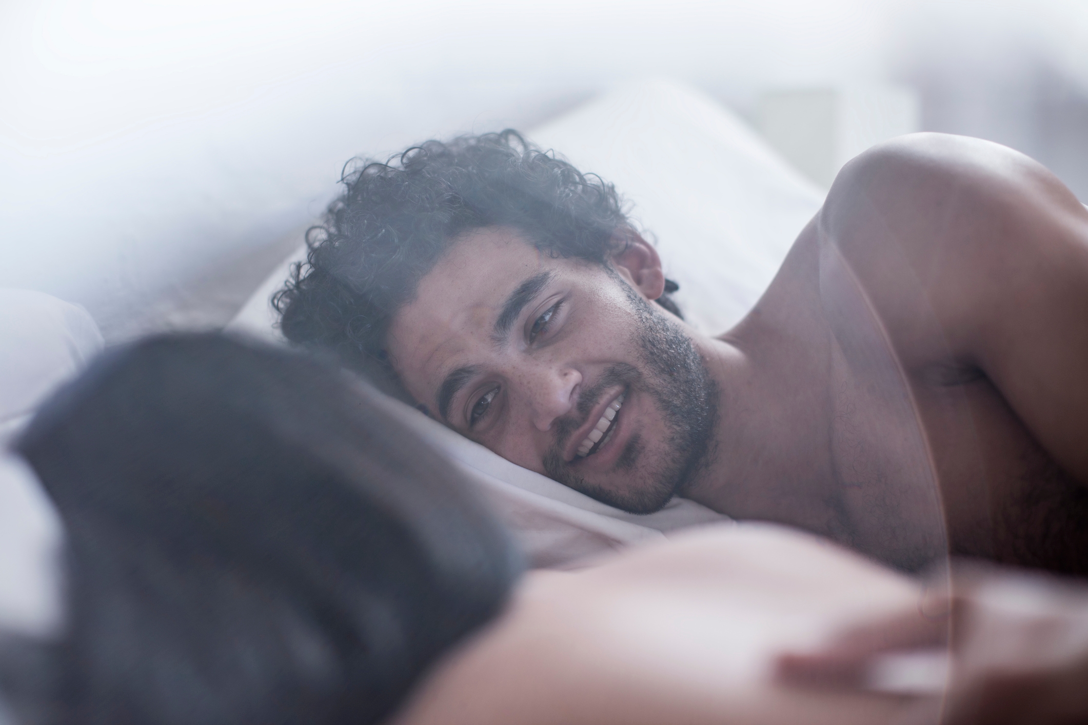 A couple relaxes in bed, smiling and making eye contact, suggesting intimacy and connection