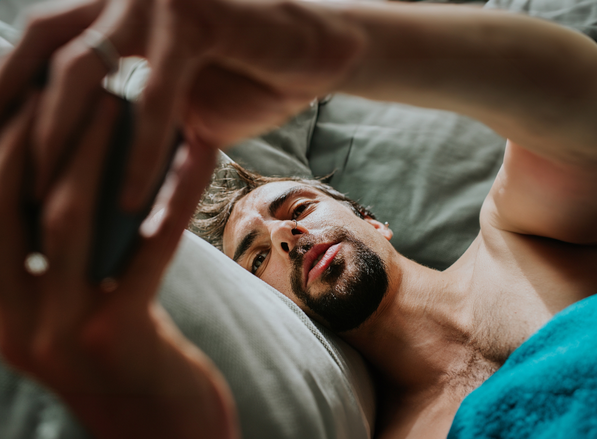Man lying in bed, holding a smartphone above him, looking at the screen intently. His expression is focused and relaxed