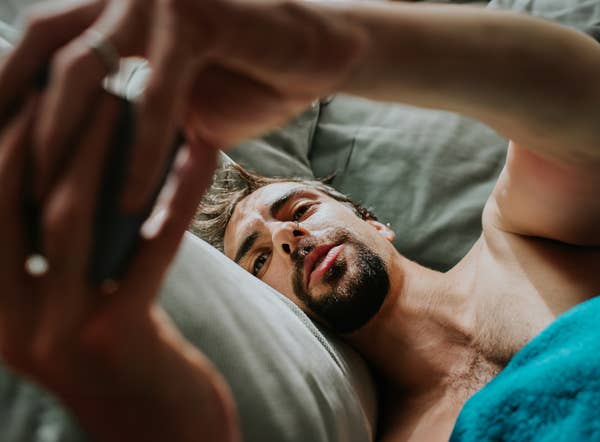 Man lying in bed, holding a smartphone above him, looking at the screen intently. His expression is focused and relaxed