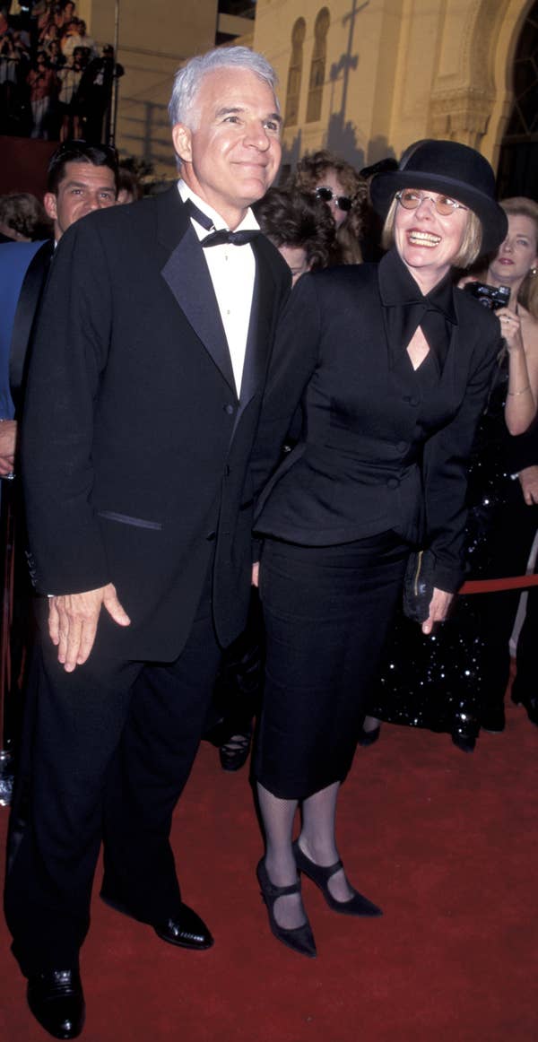 A couple in formal attire on the red carpet, the man in a tuxedo and the woman in a chic black suit with a bow tie and hat, smiling