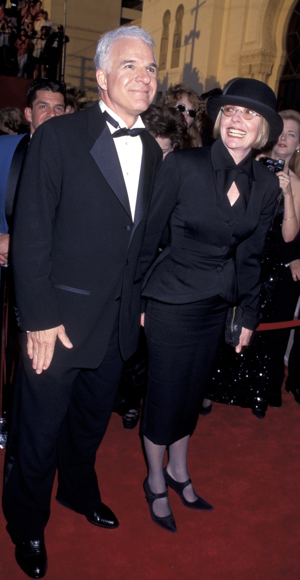 A couple in formal attire on the red carpet, the man in a tuxedo and the woman in a chic black suit with a bow tie and hat, smiling