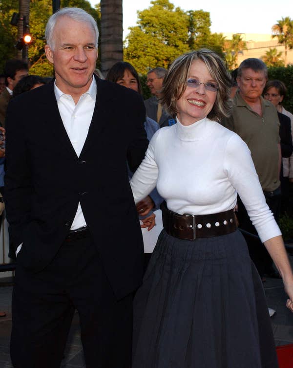 A man in a suit and a woman in a turtleneck and full skirt smile while standing outdoors at an event