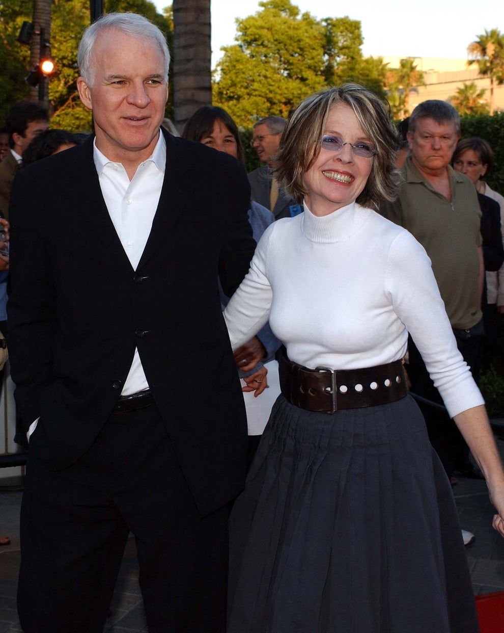 A man in a suit and a woman in a turtleneck and full skirt smile while standing outdoors at an event