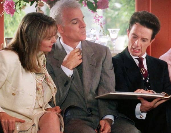 Three people in a formal setting, two in suits and one in a dress. The person on the right is holding a clipboard, appearing to explain something