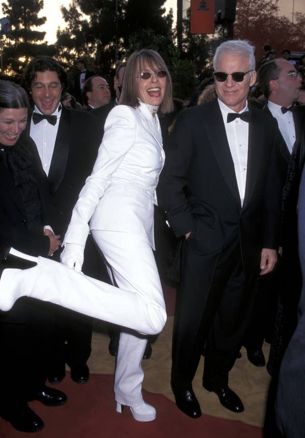 A woman in a glamorous white suit playfully kicks her leg on the red carpet. A man in a tuxedo and sunglasses stands beside her, smiling