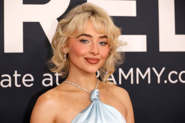 Person in elegant, halter-style gown with ruffled hair and pearl earrings, attending an event with text partially visible in the background