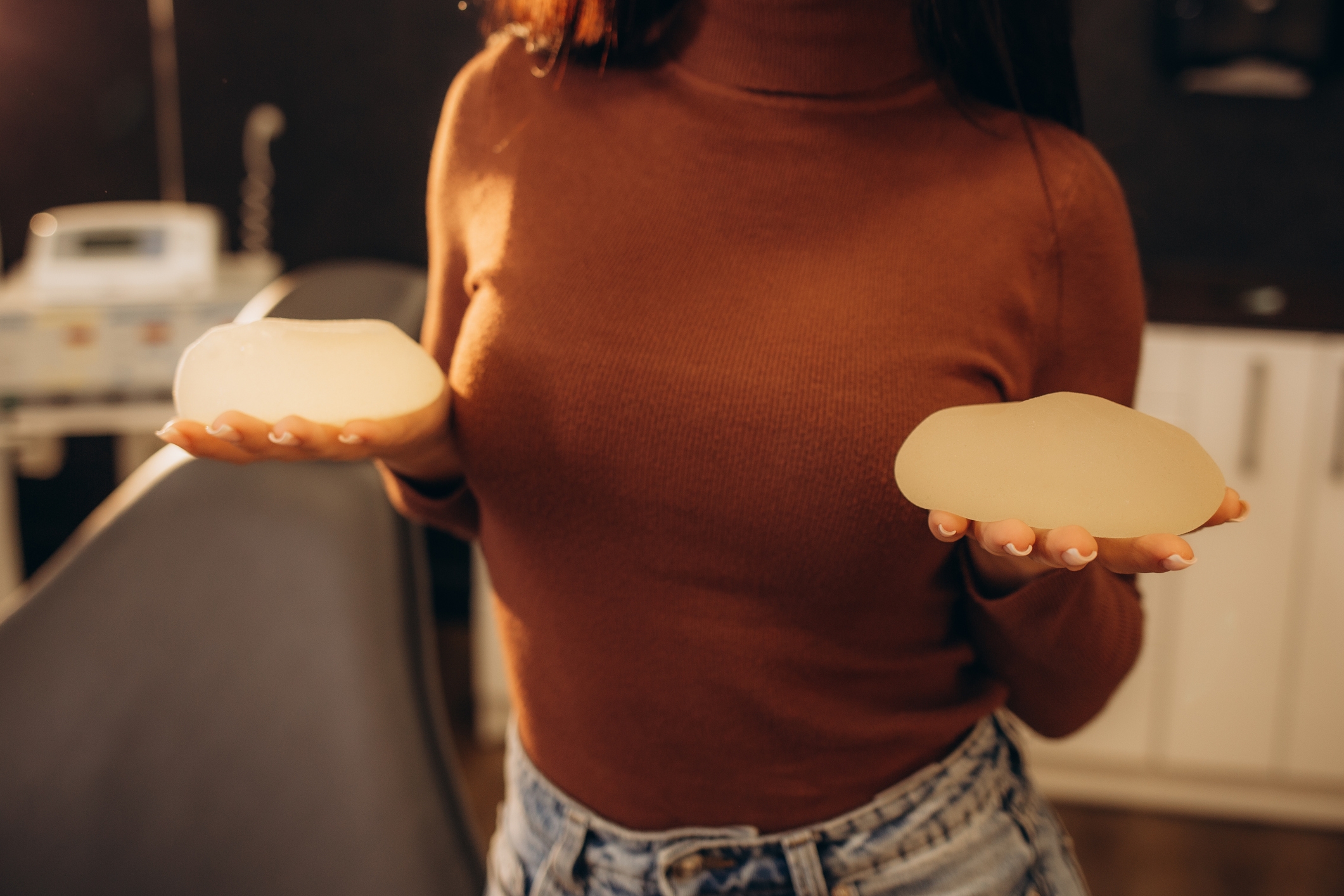 Person som holder to brystimplantater i medisinske omgivelser, iført en turtleneck og jeans