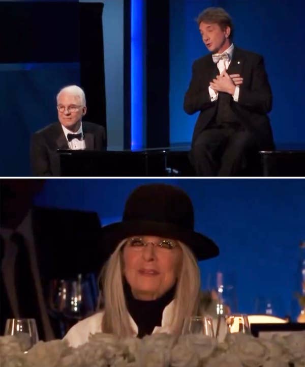 Two men in tuxedos perform on stage. A woman in a hat and glasses watches from the audience, surrounded by floral arrangements