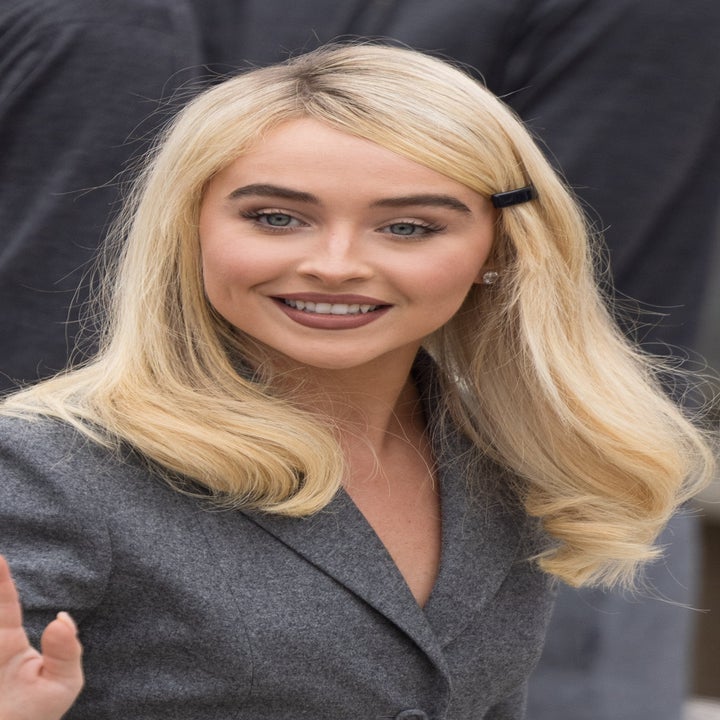 Person in a tailored, professional outfit with long sleeves and a pleated skirt waves while standing in an outdoor setting