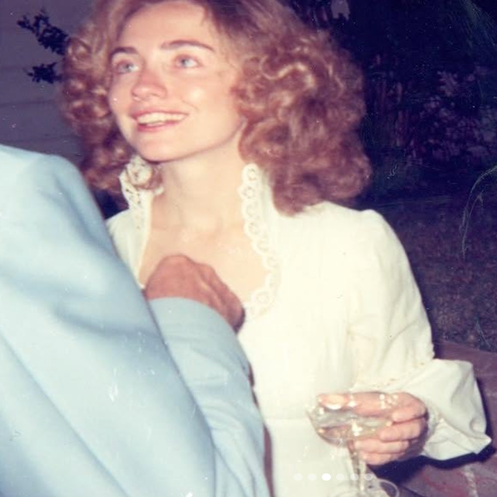 Person in vintage lace-trimmed dress holds a wine glass, smiling and looking upward, appearing engaged in a conversation