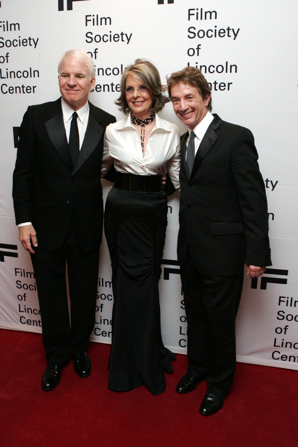Three people on the red carpet at a Film Society event, in formal attire: two men in tuxedos and a woman in a stylish gown with a fitted waist