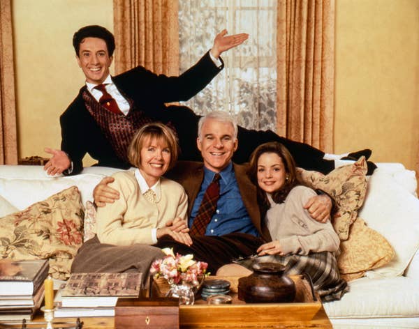 Four people smile and pose on a couch in a living room setting. They are dressed in casual, classic 1990s attire