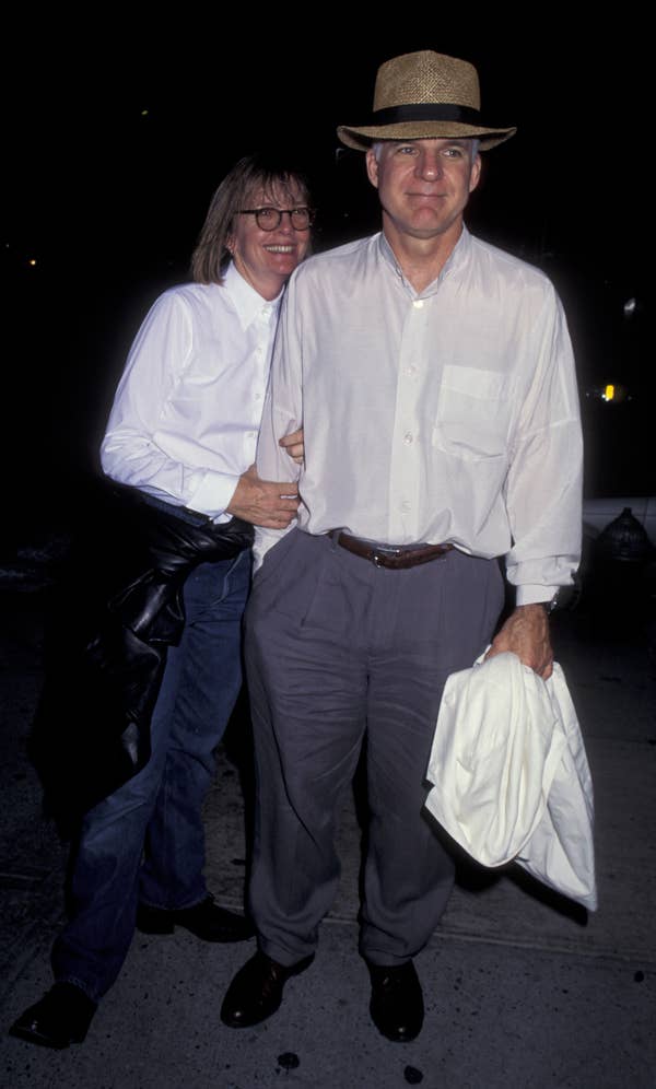 Two people pose together. One, in a hat and light shirt, holds a jacket. They're casually dressed, possibly at a celebrity event