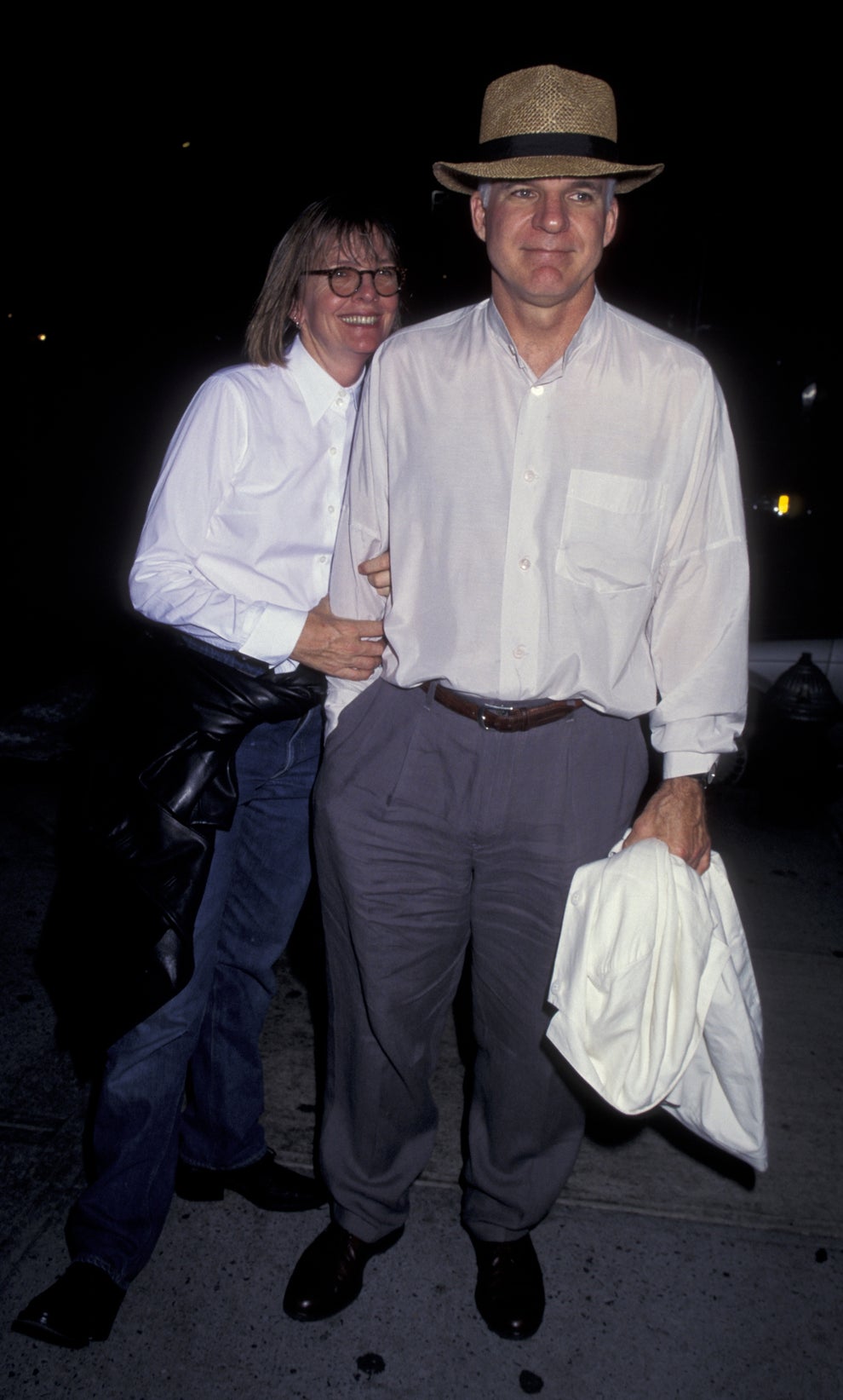 Two people pose together. One, in a hat and light shirt, holds a jacket. They're casually dressed, possibly at a celebrity event