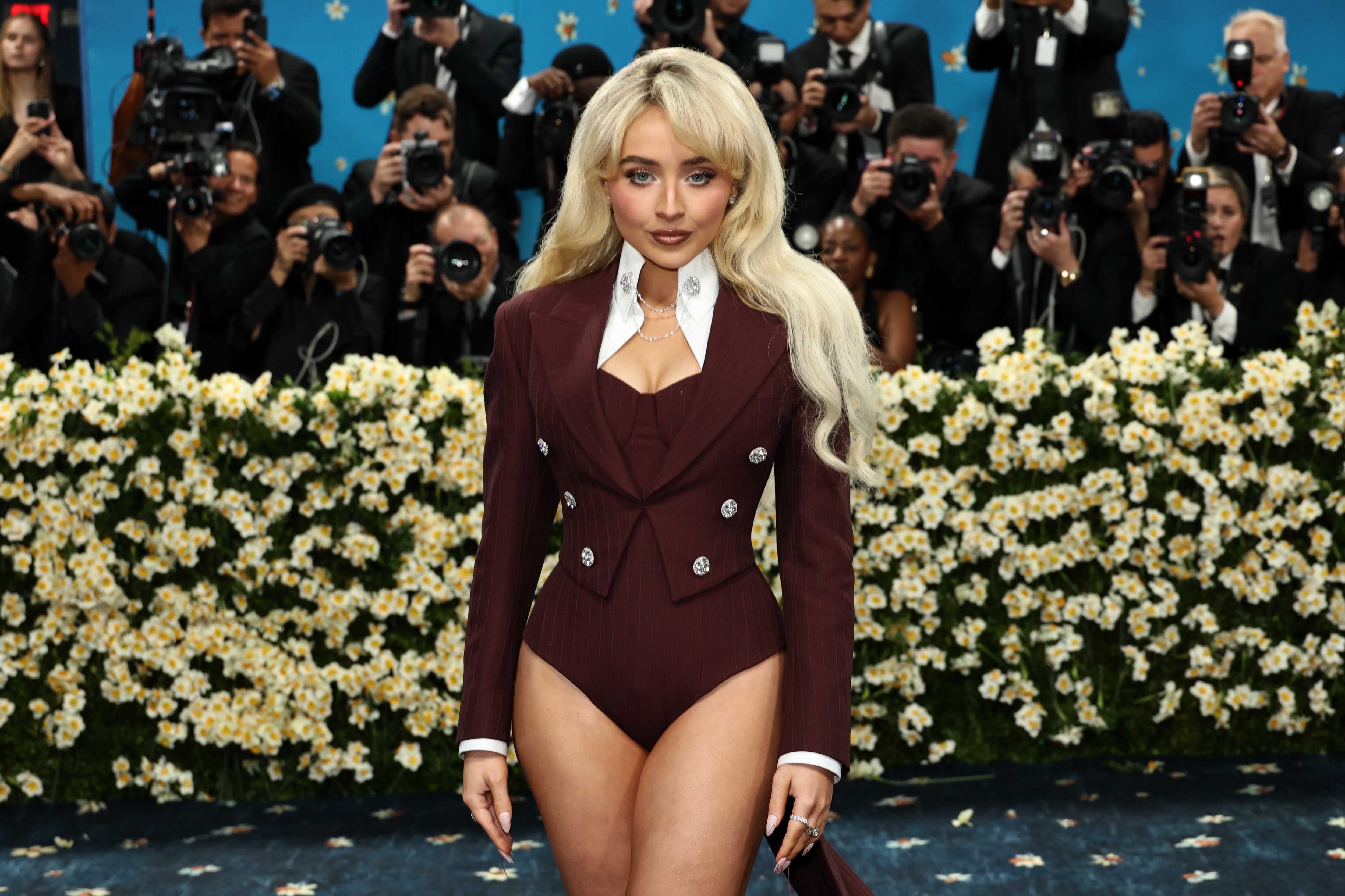 Person on red carpet in a tailored, double-breasted jacket with high-cut bottoms. Long hair, floral backdrop, photographers in the background