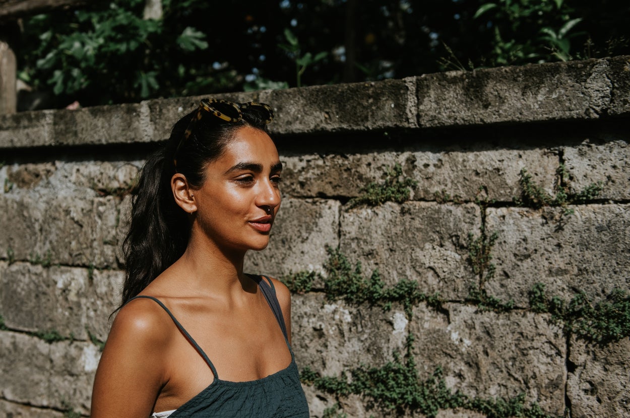 Person with long hair in a sleeveless outfit walks outside near a stone wall, looking content and relaxed
