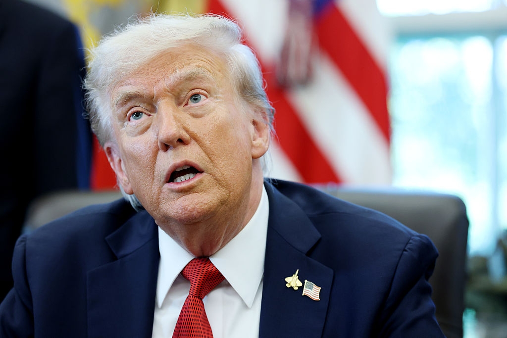 En person i dress med en amerikansk flaggnål, sitter ved et skrivebord, ser oppover, med flagg i bakgrunnen