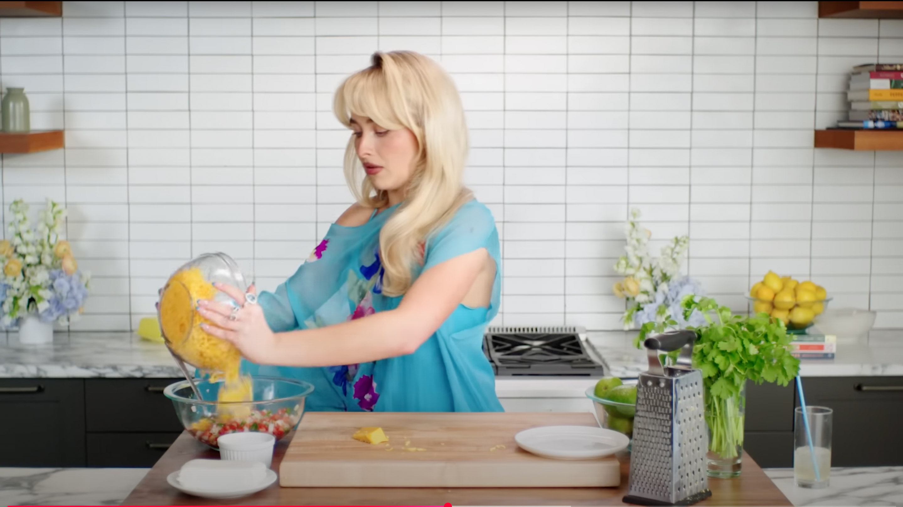 Person in a kitchen pouring blended mixture into a bowl of ingredients, with a grater, cutting board, and lemons visible