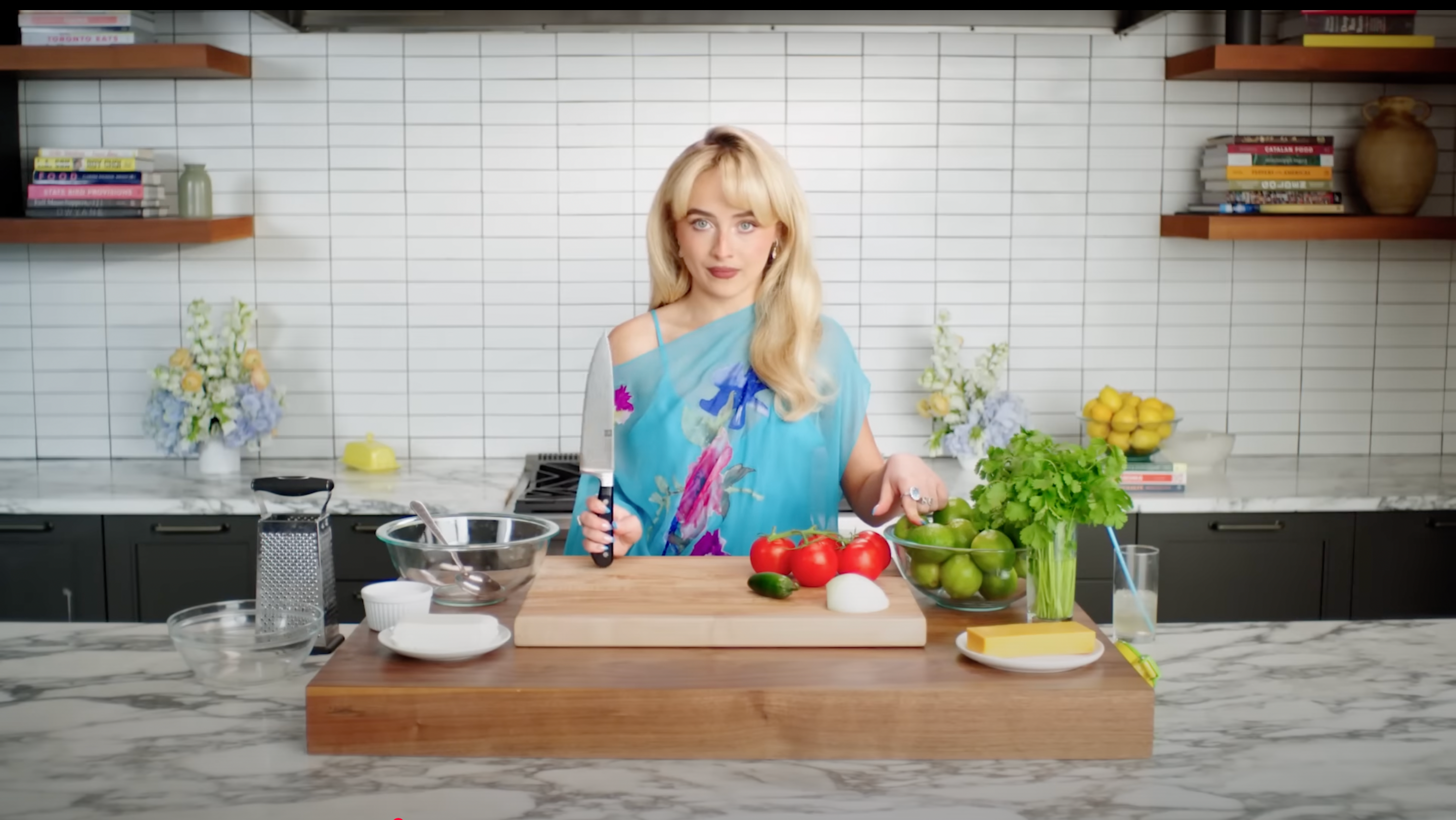 A person stands in a kitchen wearing an off-shoulder top, holding a chef's knife. Various fresh ingredients are on the counter
