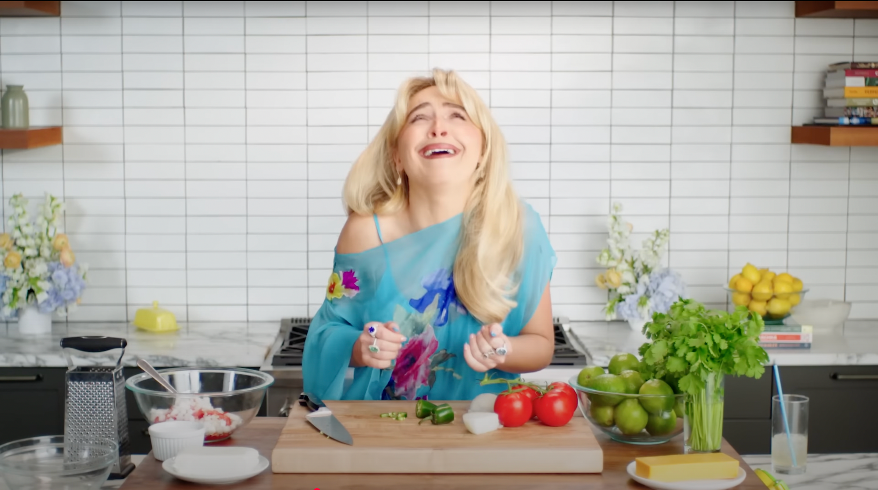 Person with long hair wearing an off-shoulder top joyfully prepares food in a modern kitchen with tomatoes, limes, and herbs on the counter