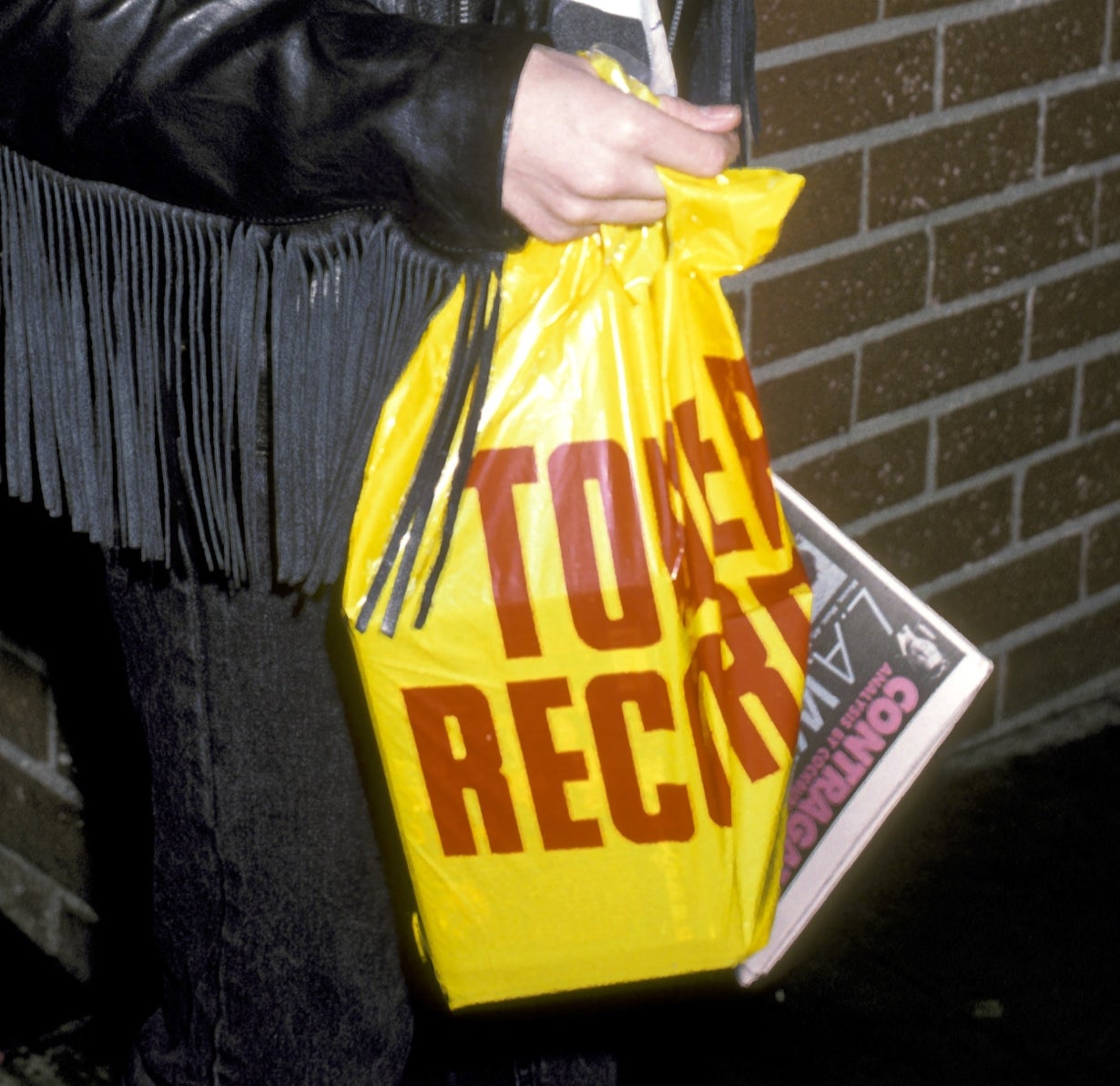 Person smiling portion holding a Tower Records bag, wearing a leather overgarment with fringes, successful beforehand of a ceramic wall
