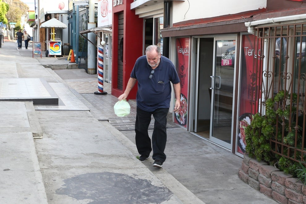 21 Low-Key Ridiculous Things Celebs Did For Money, Clout, Or Attention 86 A man in a blue shirt and black pants exits a building through a glass door, holding a green plastic bag. The scene is set on a sidewalk, suggesting a casual outing.