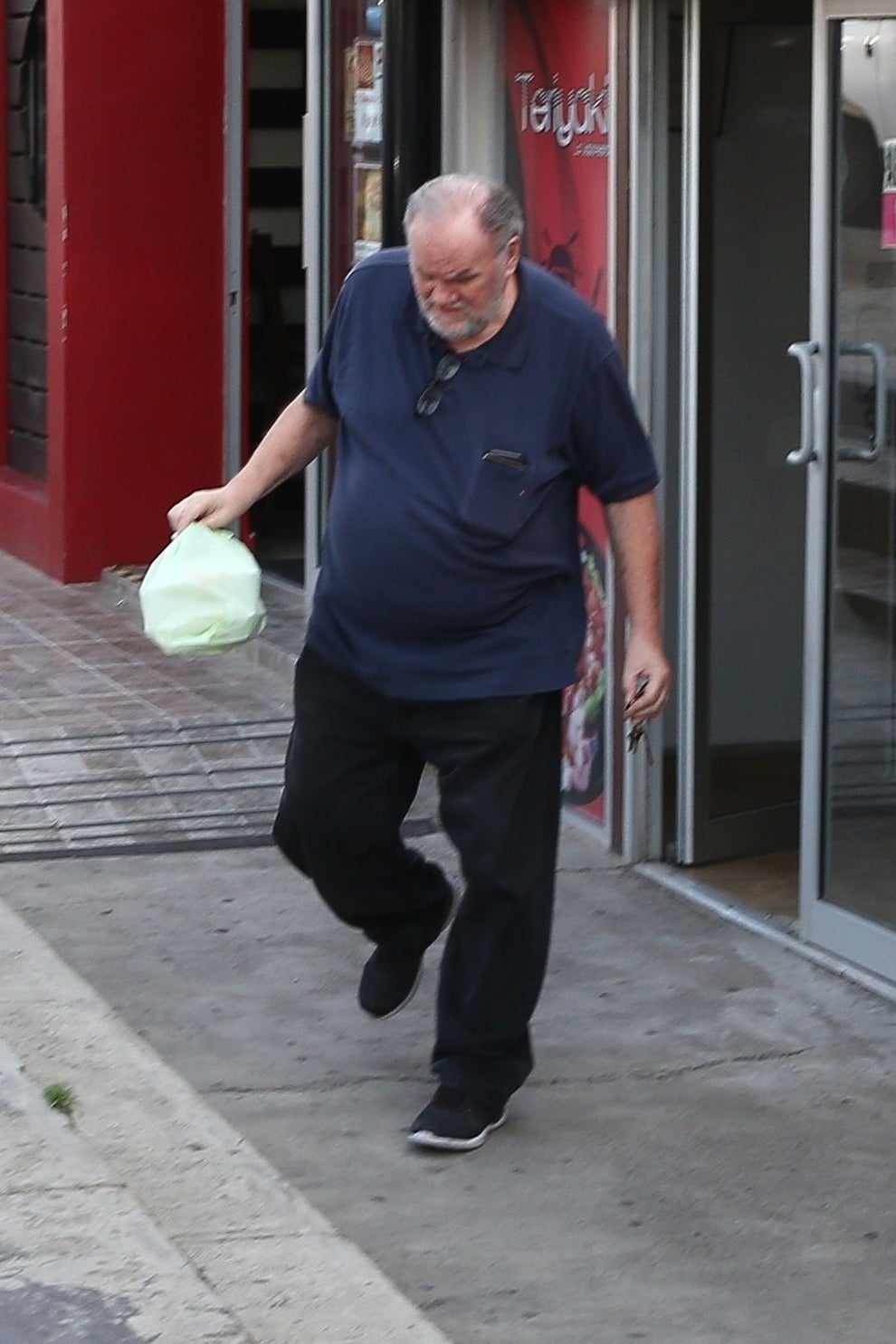 21 Low-Key Ridiculous Things Celebs Did For Money, Clout, Or Attention 80 A man in a blue shirt and black pants exits a building through a glass door, holding a green plastic bag. The scene is set on a sidewalk, suggesting a casual outing.