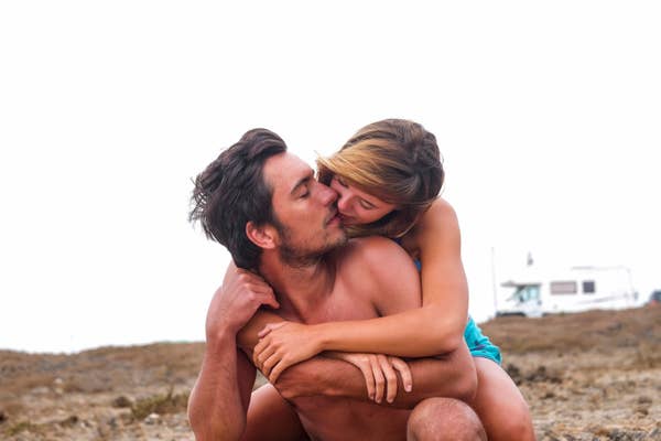 A couple embraces affectionately outdoors, with the woman hugging the man from behind and kissing his cheek