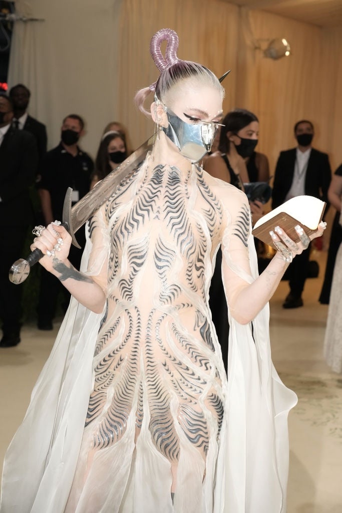 21 Low-Key Ridiculous Things Celebs Did For Money, Clout, Or Attention 4 Grimes in futuristic armor-style dress with cape, holding a sword and book, on the red carpet