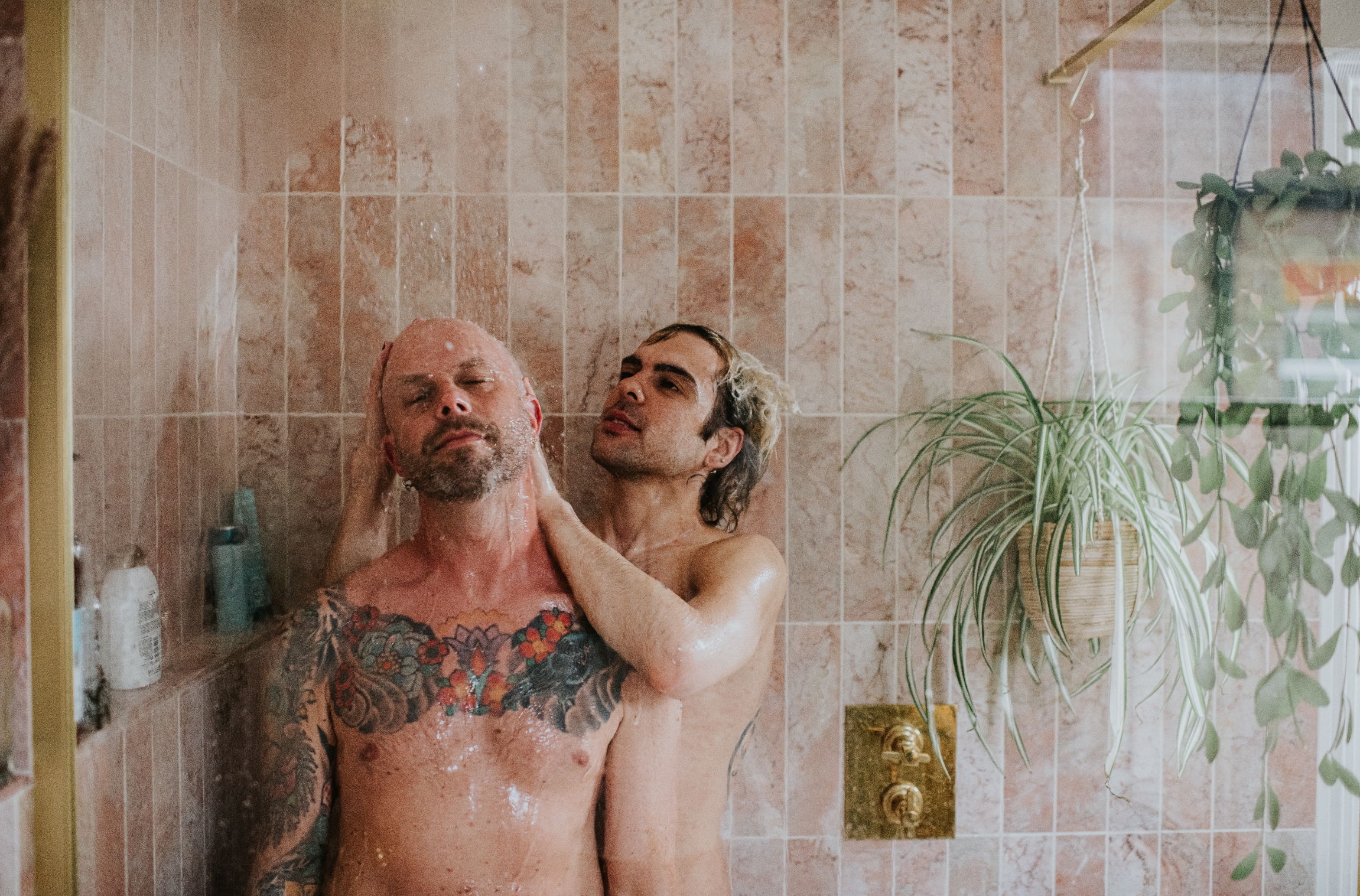 Two people in a shower share an intimate moment, with tiled walls and a hanging plant in the background