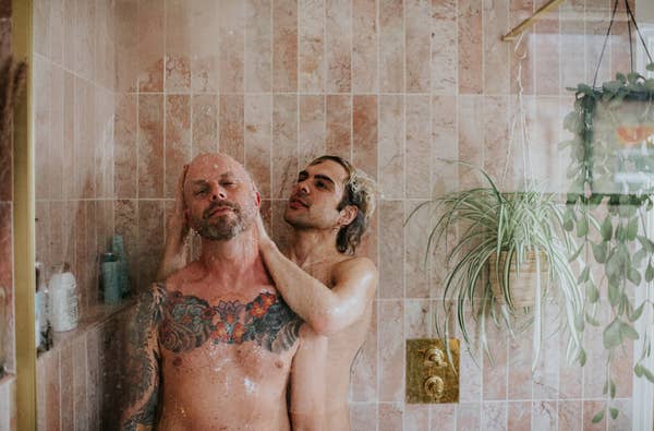 Two people in a shower share an intimate moment, with tiled walls and a hanging plant in the background