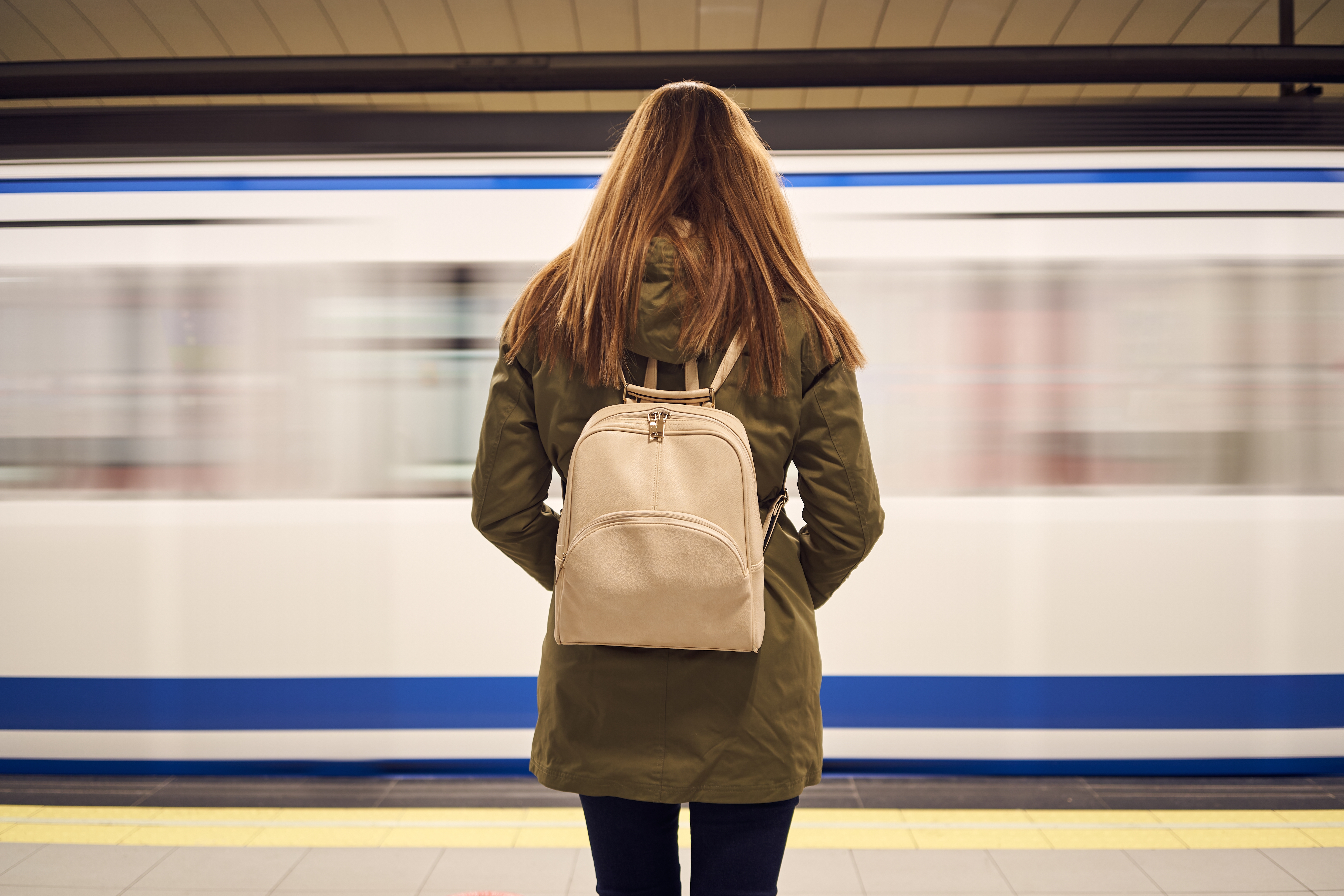 Person with a backpack waits on a subway platform as a train passes by, suggesting themes of commute and work travel