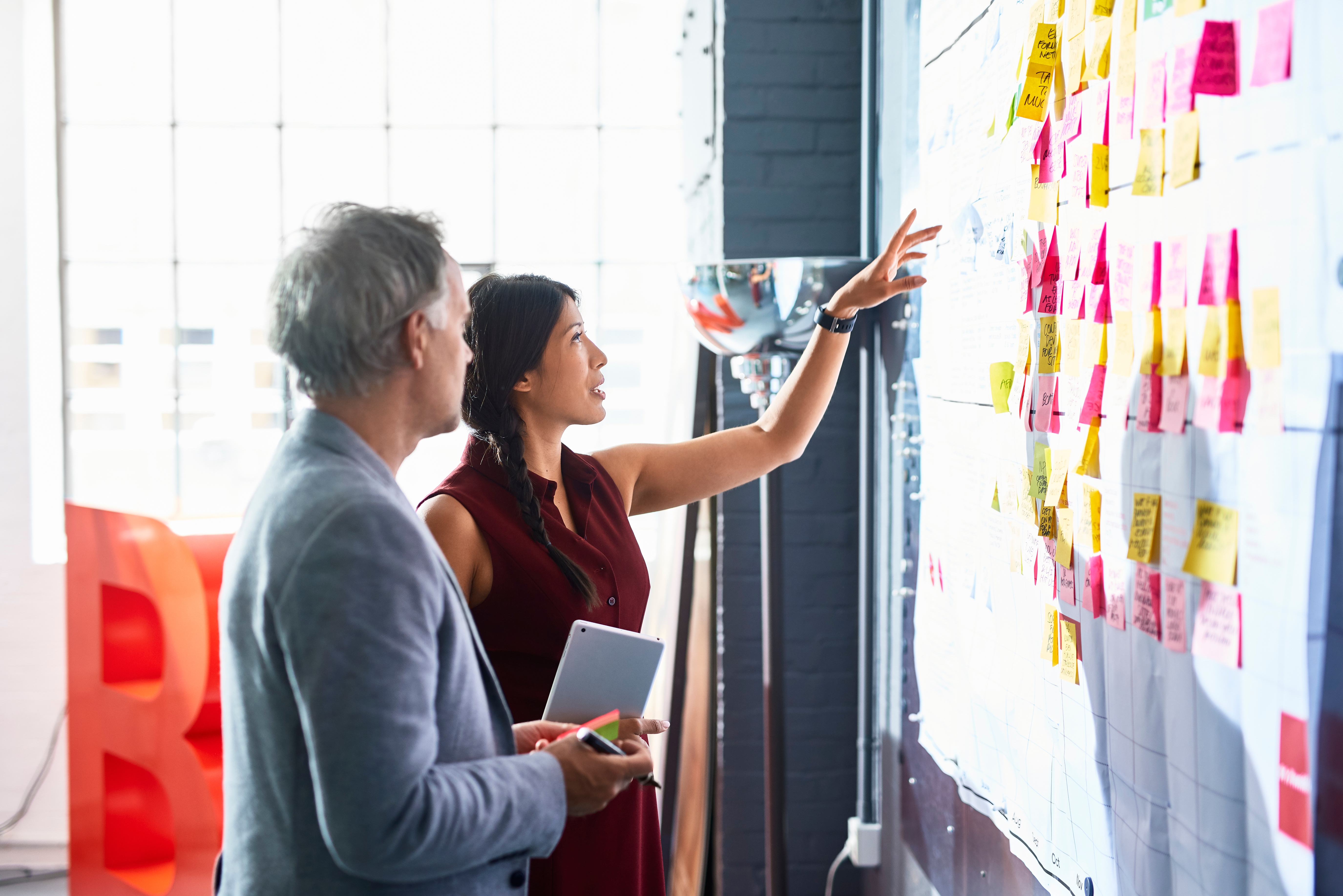 Two people stand discussing a wall covered in sticky notes. One holds a tablet, and the other points at the board, illustrating a collaborative work session