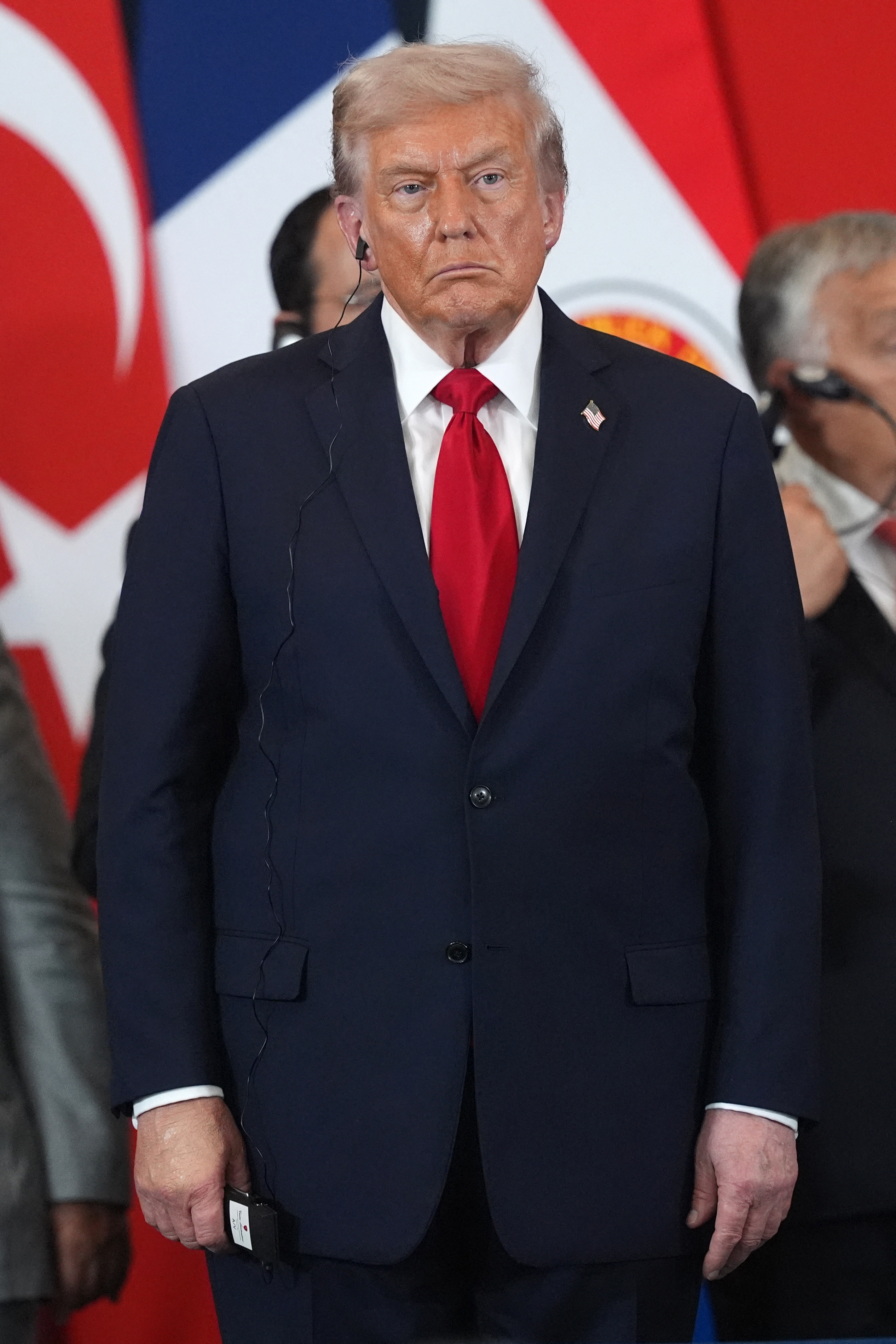 A person in a suit stands with a serious expression against a backdrop with flags, wearing an earpiece at a formal event