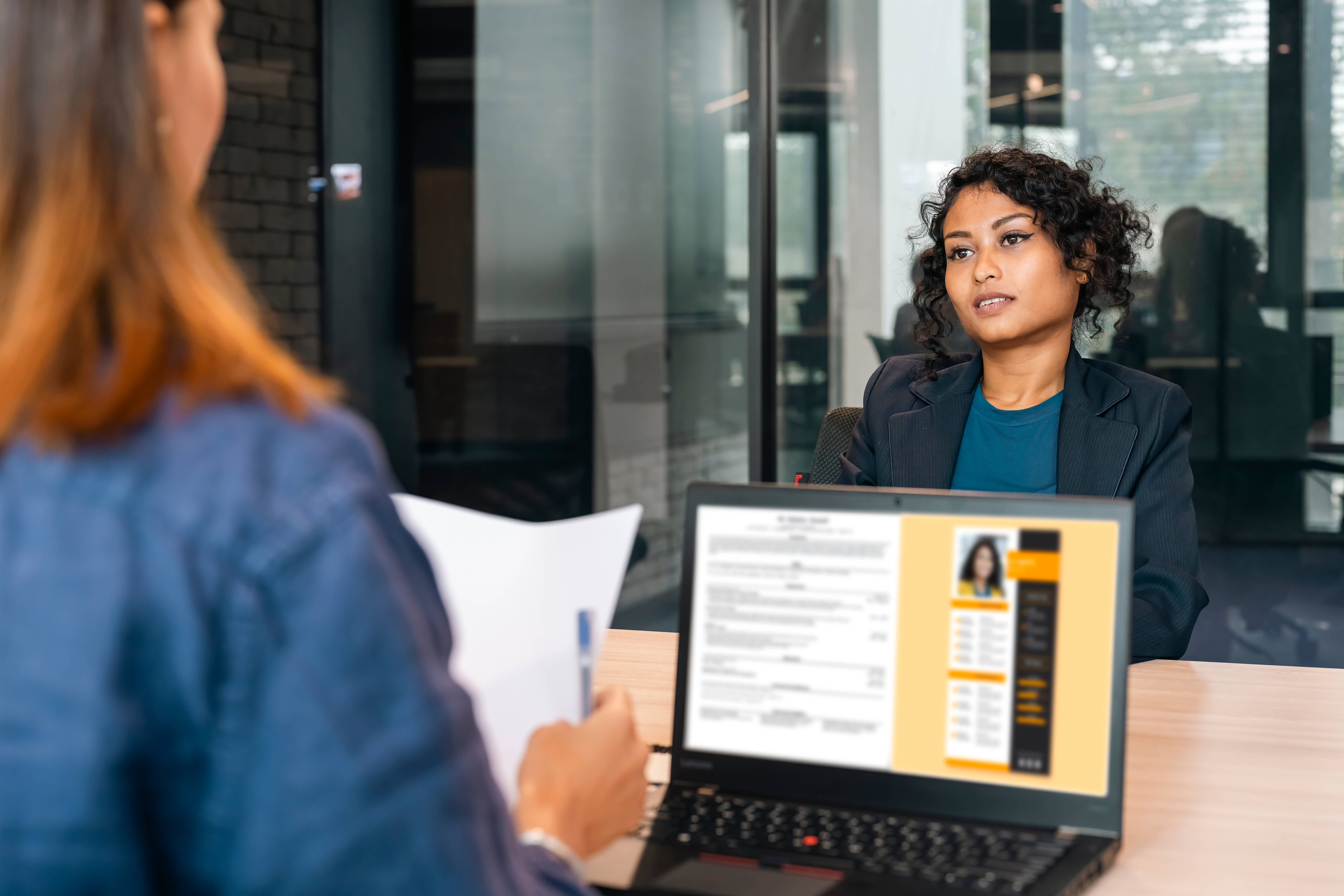 Job interview in office; woman in business attire listens attentively to interviewer with resume visible on laptop screen