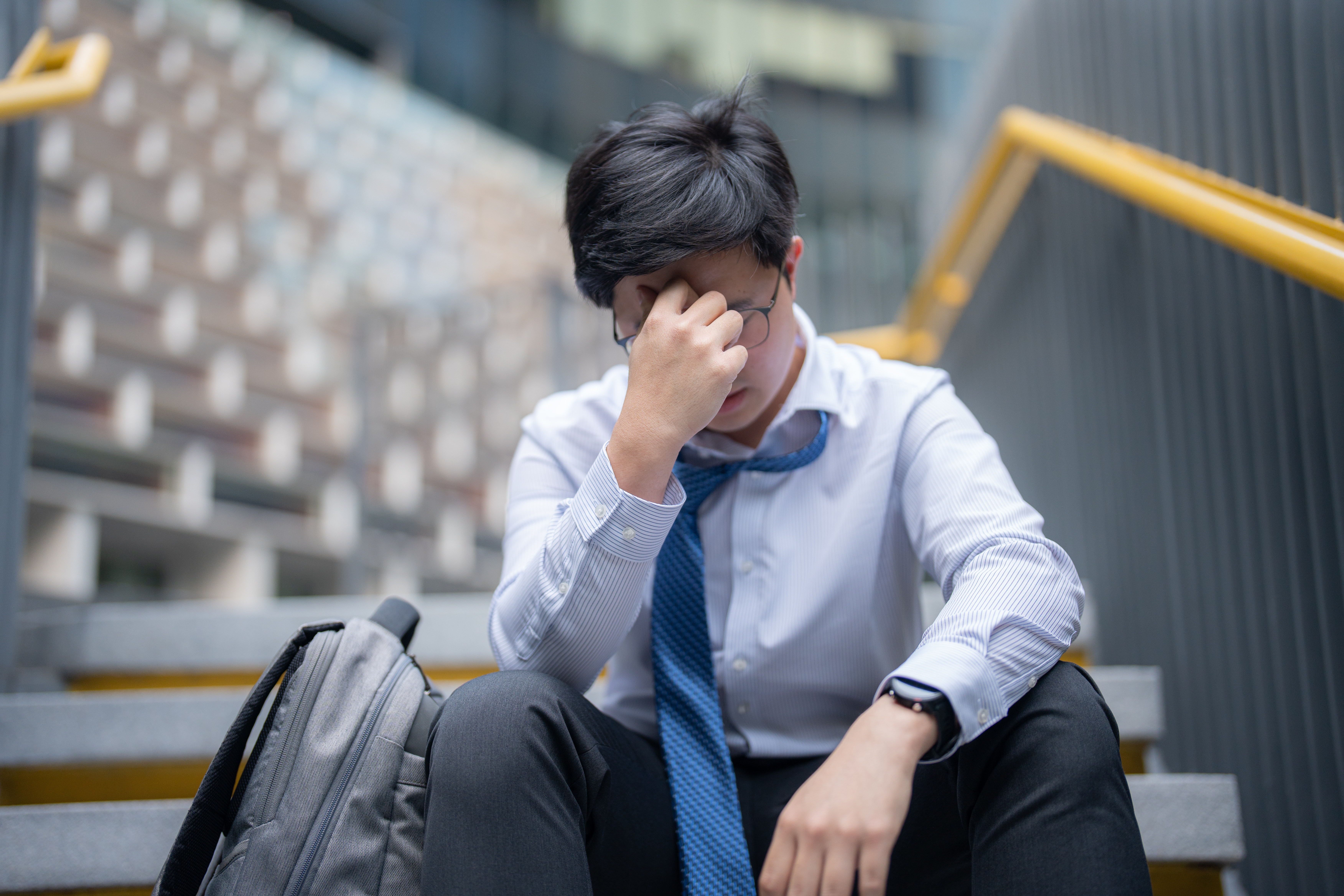 Person in an untucked shirt and loose tie, sitting on stairs with a backpack, holding their forehead in apparent stress or frustration