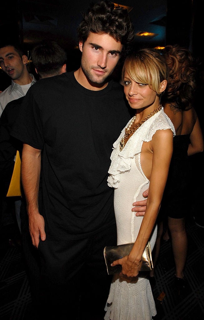 21 Low-Key Ridiculous Things Celebs Did For Money, Clout, Or Attention 34 Man and woman pose smiling at a party. The man wears a black shirt, and the woman wears a white dress, holding a gold clutch. Dim-lit, lively event.