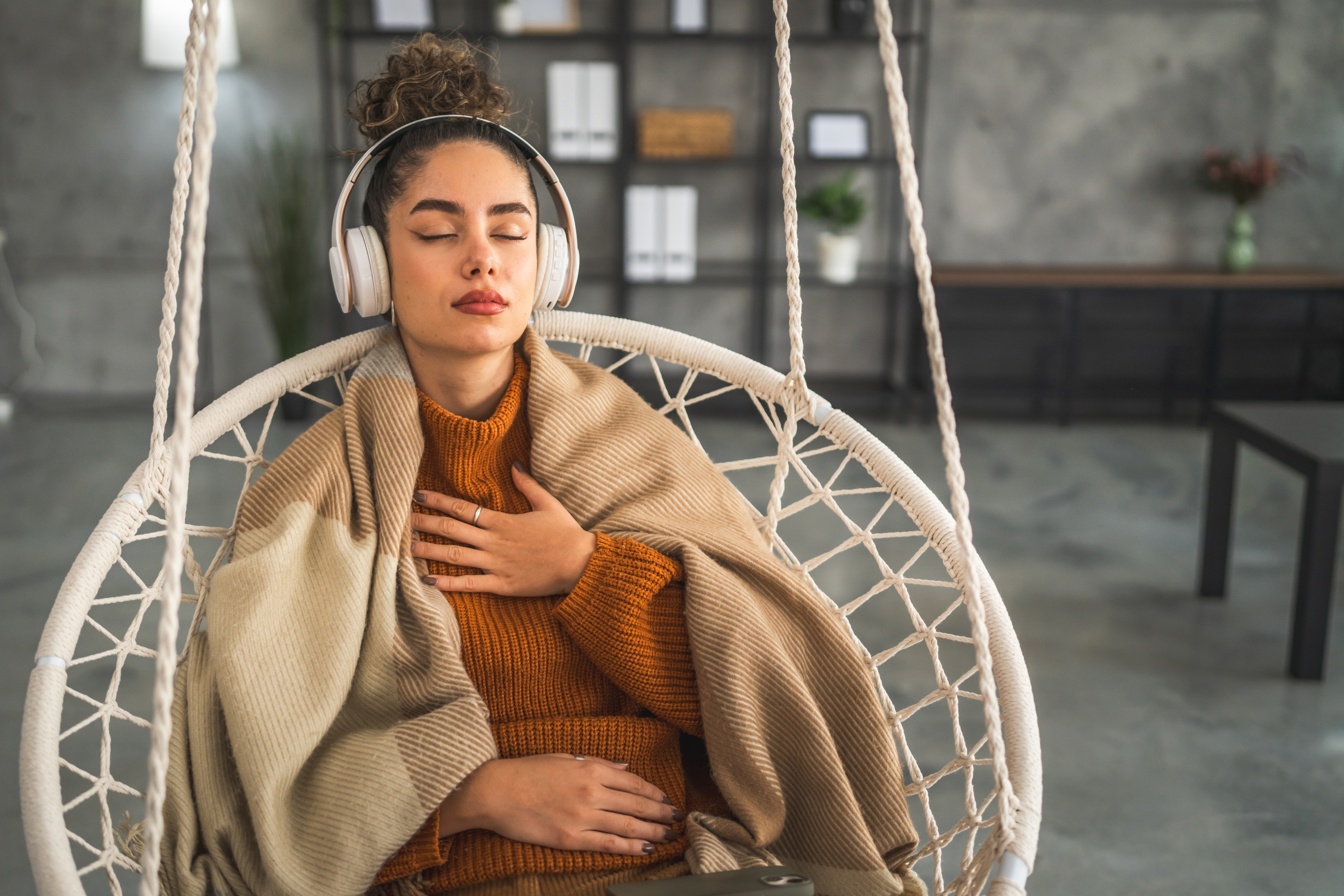 Person med hodetelefoner sitter i en hengende stol, pakket inn i et teppe, lukkede øyne, en hånd på brystet, virker avslappet