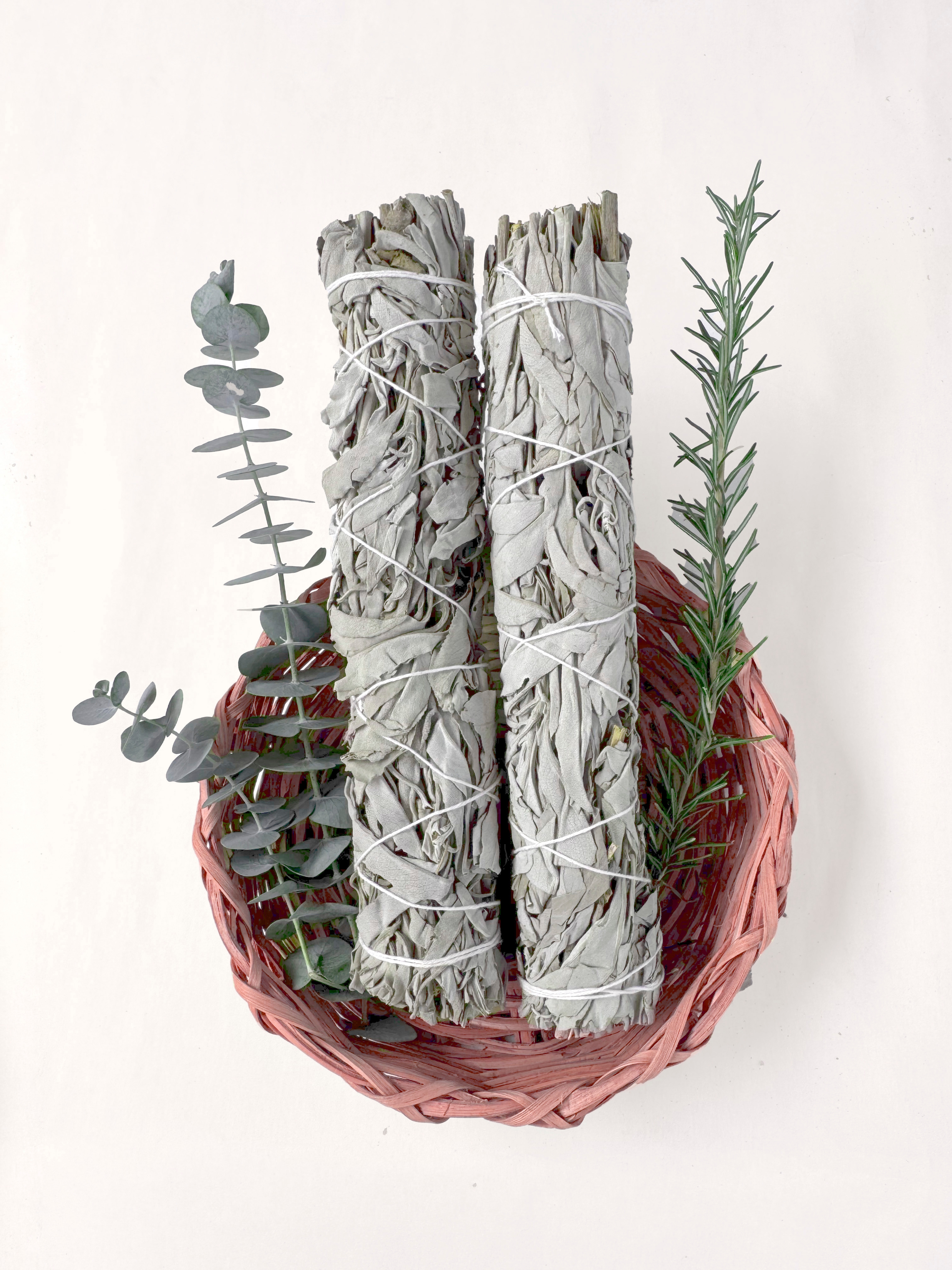 To bunter med salvie pakket inn i hvit hyssing hviler i en vevd kurv, omgitt av kvister av eukalyptus og rosmarin