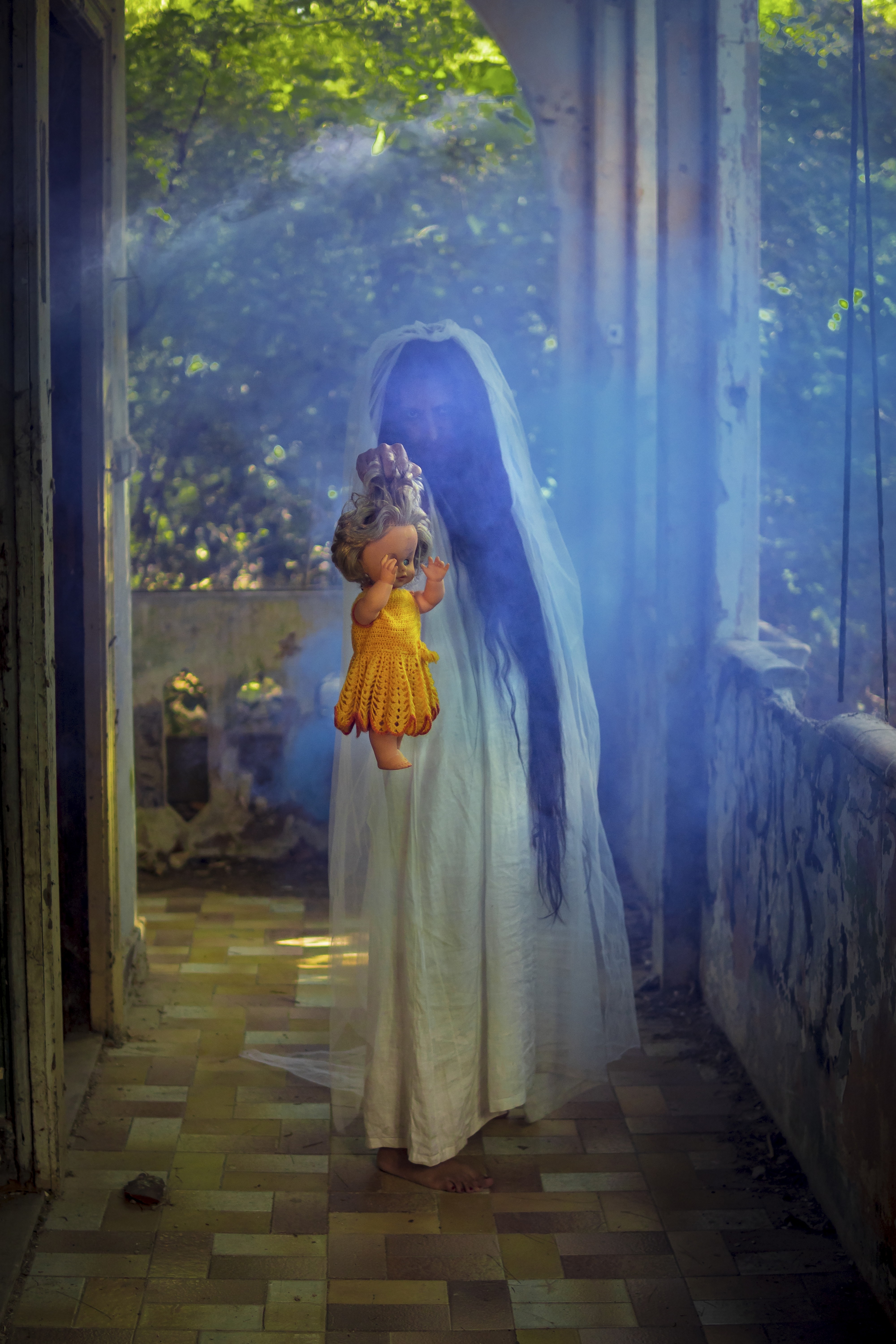 Person i en lang hvit kjole med slør, holder en dukke, står i en forvokst, forlatt setting