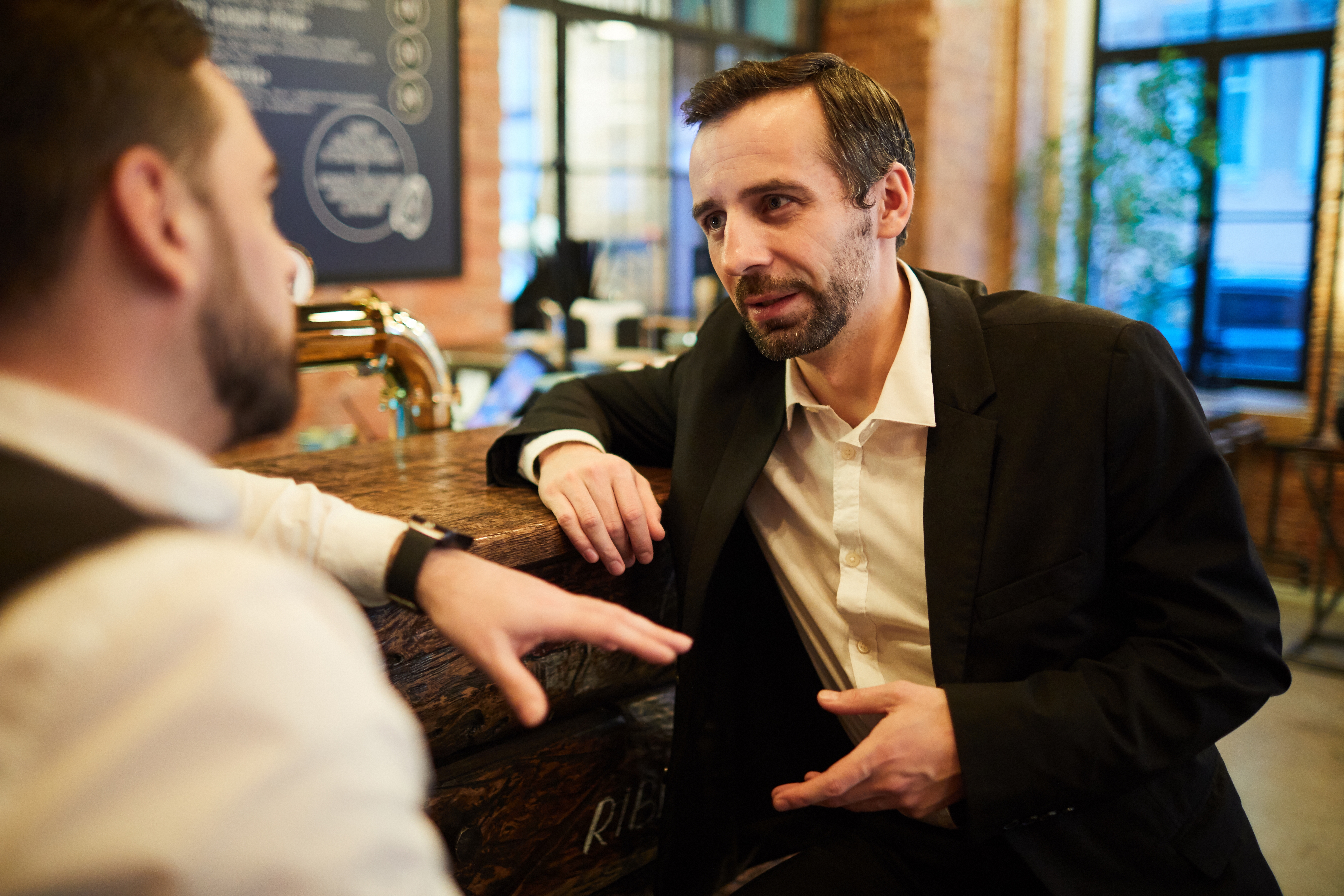 To menn er i samtale på en bar. Den ene har på seg en uformell dressjakke og skjorte, mens den andre er kledd i vest og skjorte