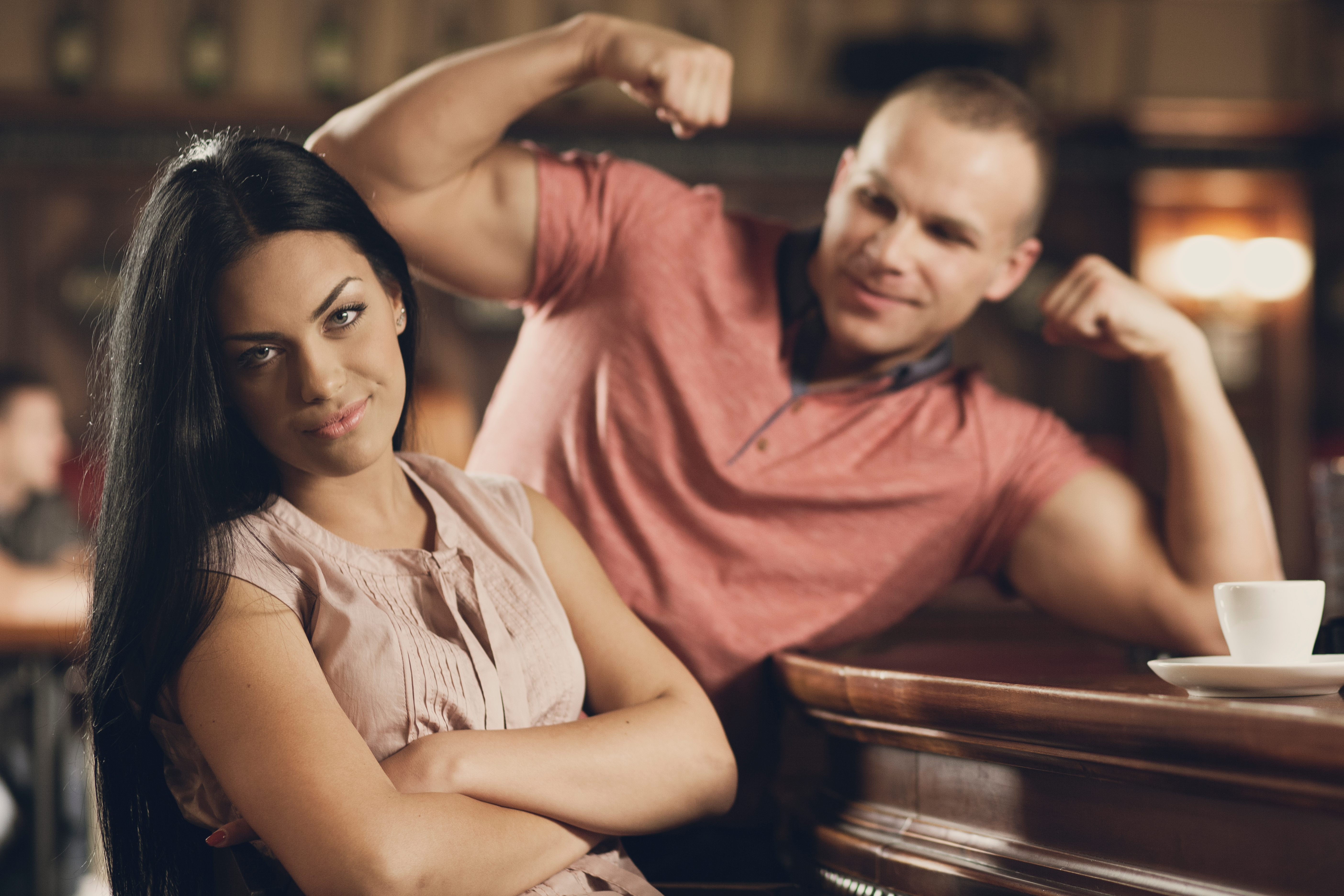 Kvinne sitter med armene i kors og ser lite imponert ut. Mann i bakgrunnen bøyer muskler, smiler. Restaurant eller kafé