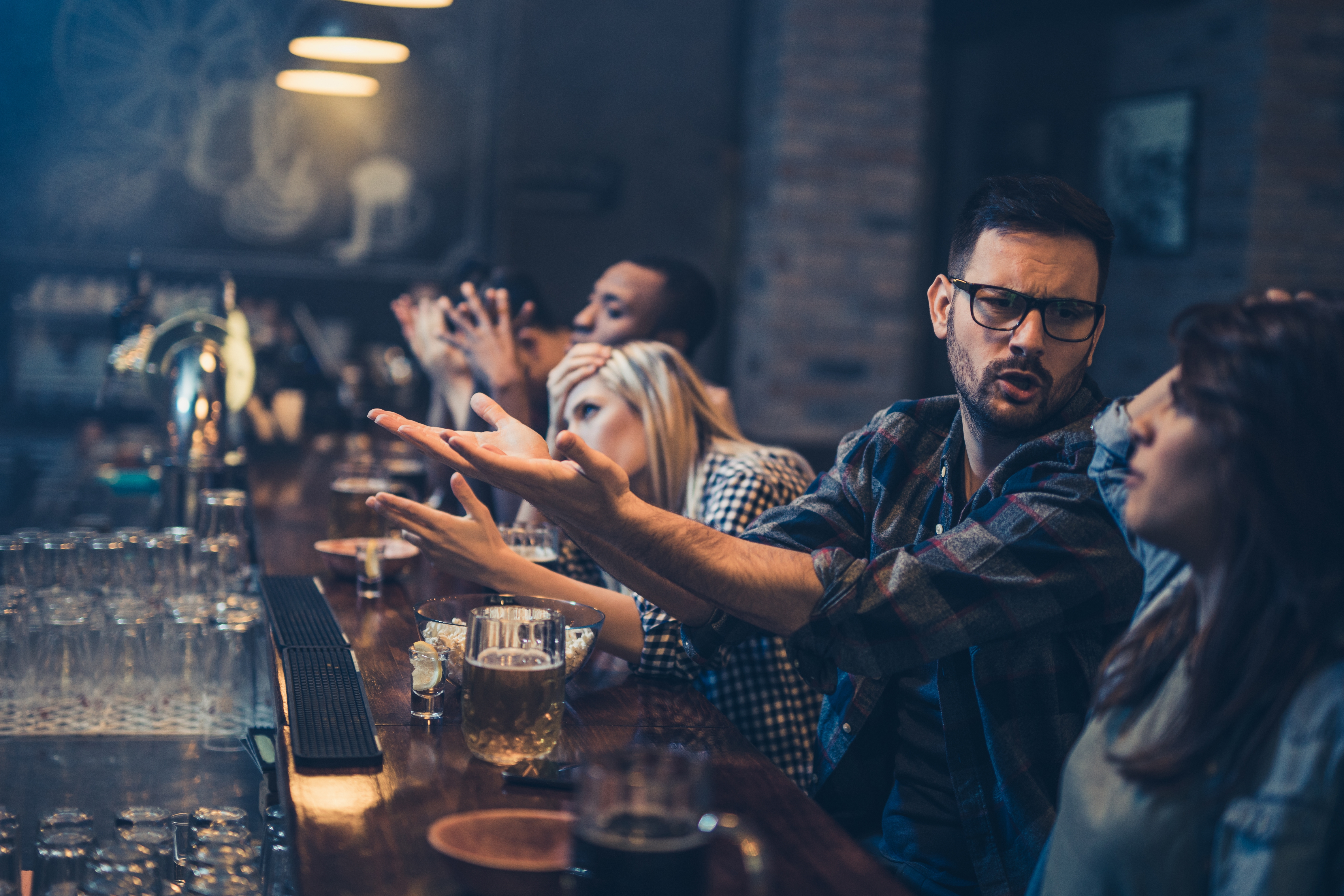 Folk er samlet på en bar og deltar i animerte samtaler. En mann gestikulerer lidenskapelig mens han snakker med en kvinne som sitter ved siden av ham